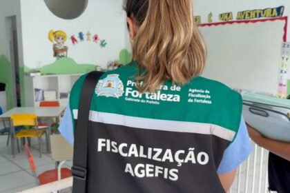 Agente da Agefis realiza fiscalização em sala de aula de escola infantil em Fortaleza, orientando sobre boas práticas sanitárias e ambientais, com mesas, cadeiras e material pedagógico ao fundo.