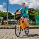 Mulher utilizando uma bicicleta compartilhada do Bicicletar em Fortaleza, pedalando em praça arborizada sob céu azul.