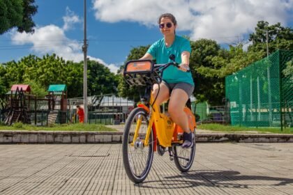 Mulher utilizando uma bicicleta compartilhada do Bicicletar em Fortaleza, pedalando em praça arborizada sob céu azul.