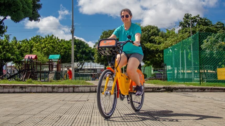 Mulher utilizando uma bicicleta compartilhada do Bicicletar em Fortaleza, pedalando em praça arborizada sob céu azul.