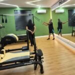 Grupo de idosos realiza exercícios de pilates em sala equipada do Centro Dia do Idoso de Eusébio, com aparelhos, espelhos e instrutor orientando a atividade.