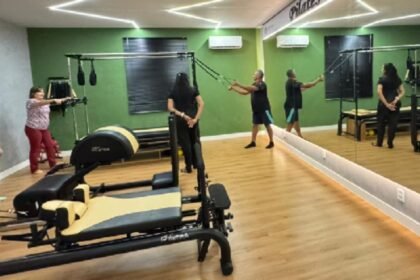 Grupo de idosos realiza exercícios de pilates em sala equipada do Centro Dia do Idoso de Eusébio, com aparelhos, espelhos e instrutor orientando a atividade.