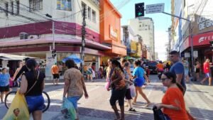 Pessoas atravessam rua comercial movimentada no Centro de Fortaleza, com lojas abertas, pedestres carregando sacolas e tráfego urbano ao fundo.