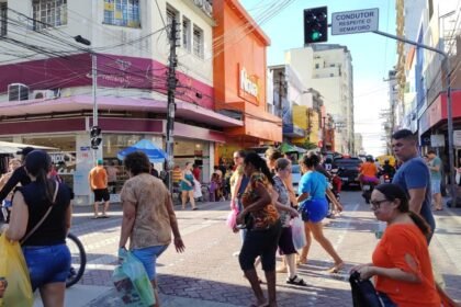 Pessoas atravessam rua comercial movimentada no Centro de Fortaleza, com lojas abertas, pedestres carregando sacolas e tráfego urbano ao fundo.