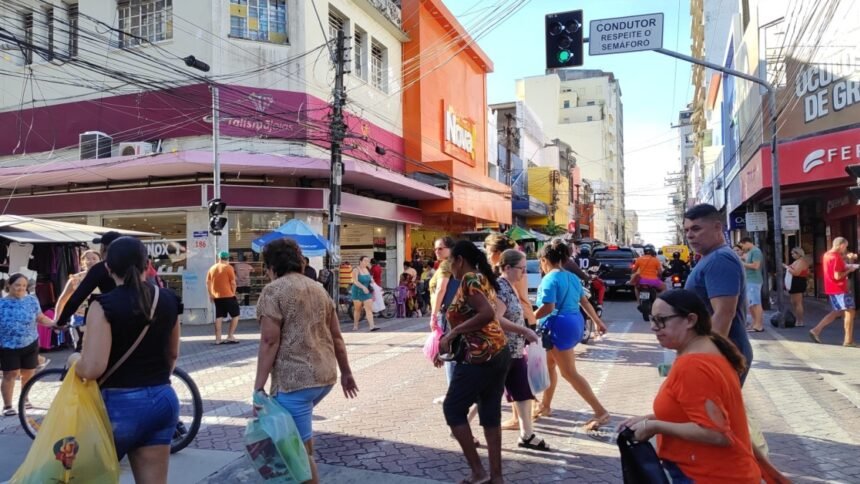 Pessoas atravessam rua comercial movimentada no Centro de Fortaleza, com lojas abertas, pedestres carregando sacolas e tráfego urbano ao fundo.