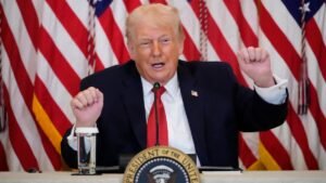 Donald Trump fala ao microfone em mesa oficial da Casa Branca, com bandeiras dos Estados Unidos ao fundo, gesticulando com as mãos durante discurso.