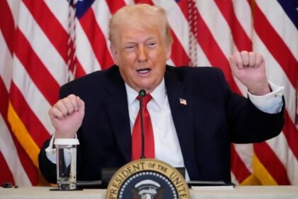 Donald Trump fala ao microfone em mesa oficial da Casa Branca, com bandeiras dos Estados Unidos ao fundo, gesticulando com as mãos durante discurso.