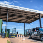 Fachada do Complexo Penitenciário Estadual de Itaitinga II, com guarita, cones de sinalização e viatura do GAP estacionada sob o pórtico de entrada, em dia ensolarado no Ceará.