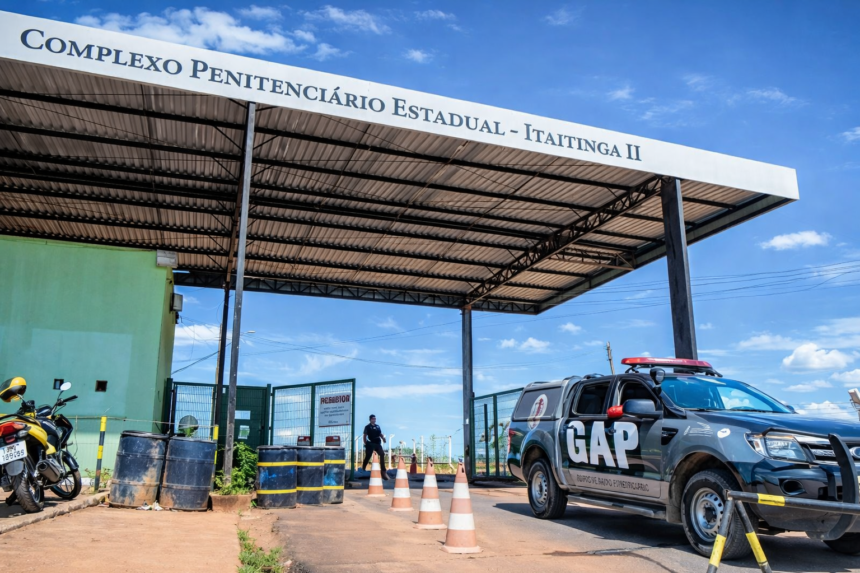 Fachada do Complexo Penitenciário Estadual de Itaitinga II, com guarita, cones de sinalização e viatura do GAP estacionada sob o pórtico de entrada, em dia ensolarado no Ceará.