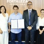 Autoridades municipais e representantes do Governo Federal posam durante a assinatura do termo de adesão de Fortaleza ao Programa de Privacidade e Segurança da Informação, com o prefeito segurando o documento oficial no auditório da Prefeitura.