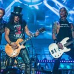 Integrantes da banda Guns N’ Roses se apresentam em palco iluminado, com guitarras e baixo em destaque, durante show com grande estrutura de luz e telões ao fundo.