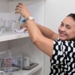 Mulher limpa prateleira com copos e utensílios em ambiente doméstico, representando o trabalho de profissionais autônomos de serviços residenciais.