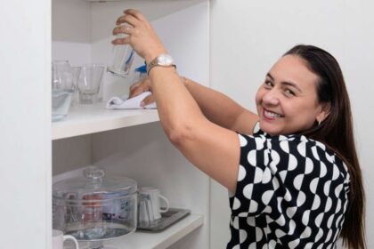 Mulher limpa prateleira com copos e utensílios em ambiente doméstico, representando o trabalho de profissionais autônomos de serviços residenciais.