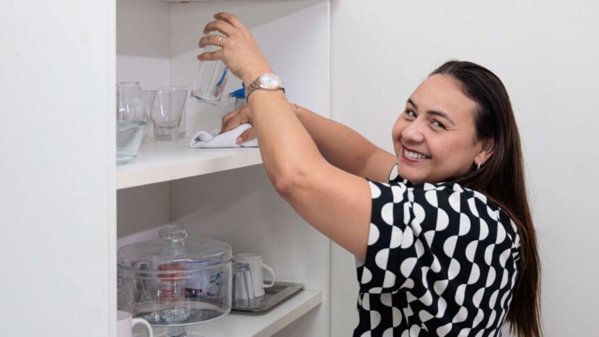 Mulher limpa prateleira com copos e utensílios em ambiente doméstico, representando o trabalho de profissionais autônomos de serviços residenciais.