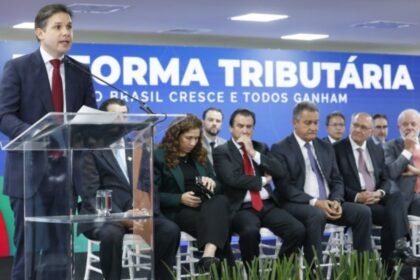 Hugo Motta fala ao microfone em evento sobre a reforma tributária, com autoridades sentadas ao fundo diante de painel com a frase “Reforma Tributária – O Brasil cresce e todos ganham”.
