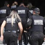 Agentes do Gaeco sobem escadaria de prédio durante operação, vestindo coletes pretos com identificação do grupo de combate às organizações criminosas.