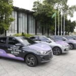 Fila de carros identificados como viaturas da Patrulha Maria da Penha estacionados em área externa, com prédios ao fundo, prontos para distribuição aos municípios do Ceará.