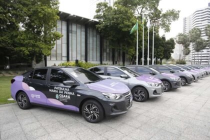 Fila de carros identificados como viaturas da Patrulha Maria da Penha estacionados em área externa, com prédios ao fundo, prontos para distribuição aos municípios do Ceará.