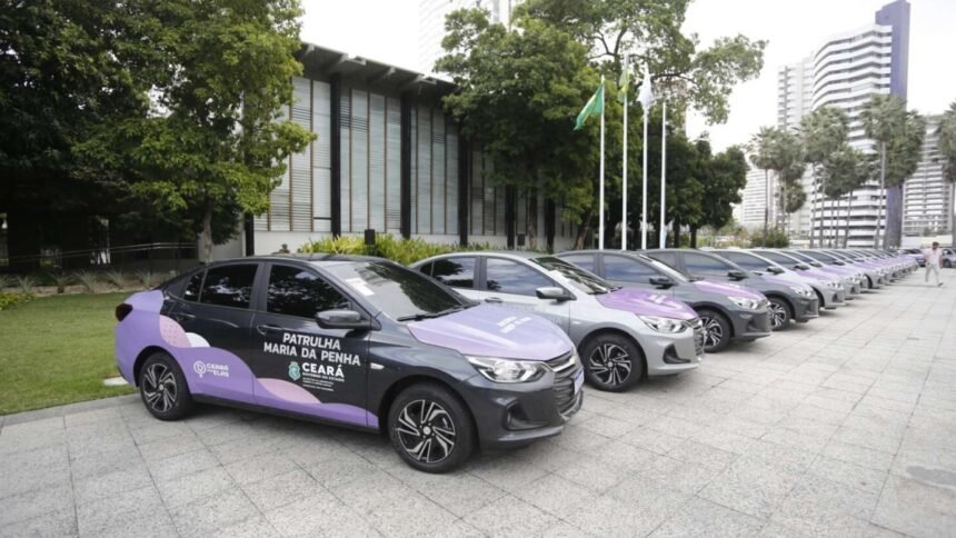Fila de carros identificados como viaturas da Patrulha Maria da Penha estacionados em área externa, com prédios ao fundo, prontos para distribuição aos municípios do Ceará.