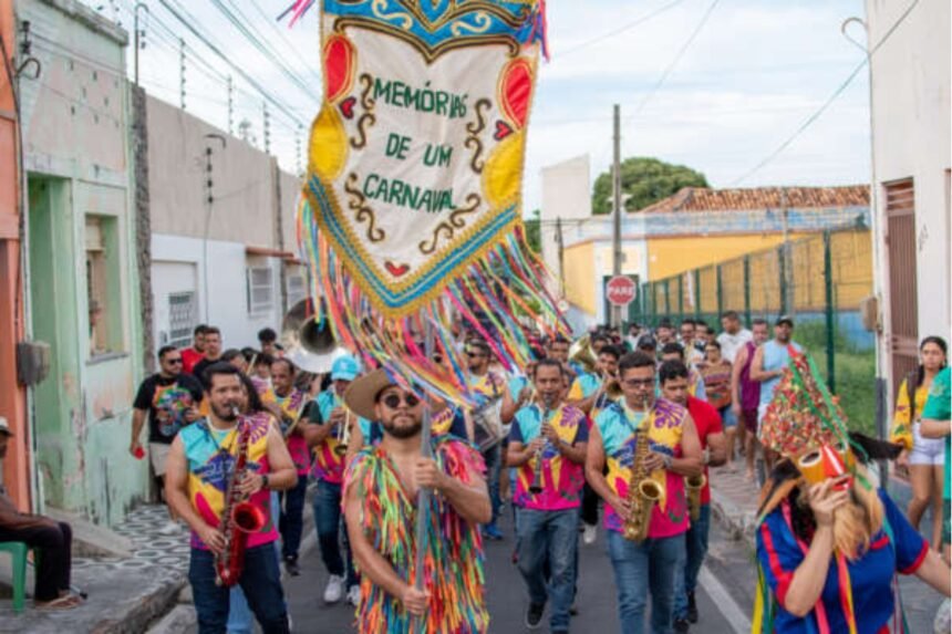 Bloco carnavalesco com músicos e foliões desfila por rua do Centro Histórico de Juazeiro do Norte, carregando estandarte “Memórias de um Carnaval” e tocando instrumentos de sopro.