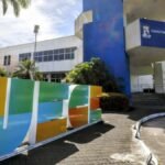 Fachada da Reitoria da Universidade Estadual do Ceará, em Fortaleza, com prédio branco e azul, palmeiras ao redor e o nome “UECE” em destaque na área externa.