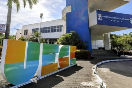 Fachada da Reitoria da Universidade Estadual do Ceará, em Fortaleza, com prédio branco e azul, palmeiras ao redor e o nome “UECE” em destaque na área externa.