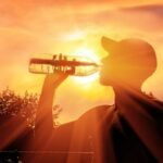 Silhueta de uma pessoa bebendo água sob o sol forte ao entardecer, com luz intensa e céu alaranjado, simbolizando o aumento das temperaturas e os efeitos do calor extremo.