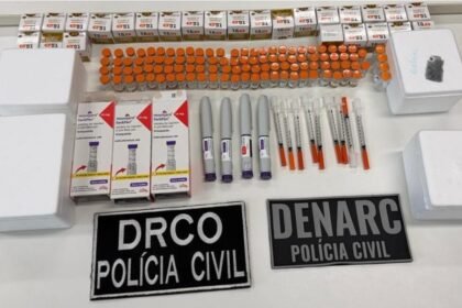 Mesa com dezenas de ampolas, caixas e canetas de medicamentos apreendidos pela Polícia Civil, além de seringas e embalagens, exibidos ao lado de placas da DRCO e da Denarc.