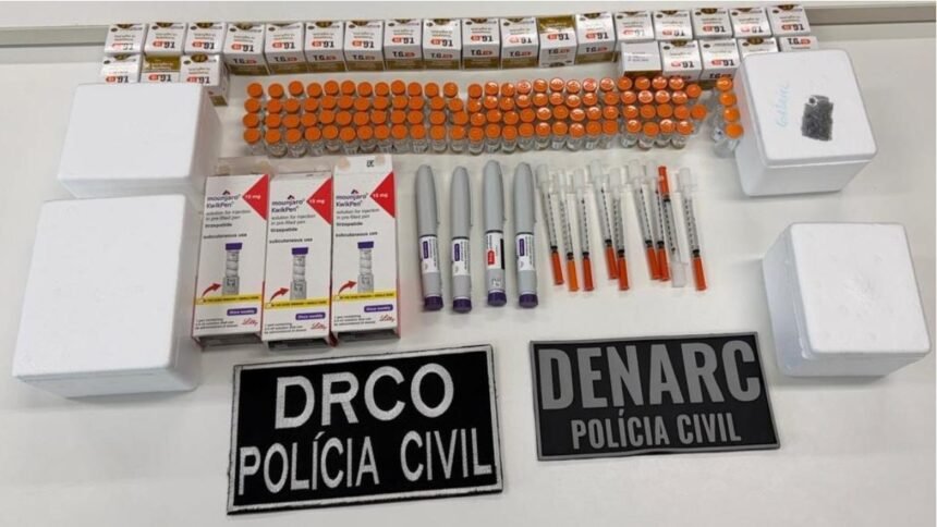 Mesa com dezenas de ampolas, caixas e canetas de medicamentos apreendidos pela Polícia Civil, além de seringas e embalagens, exibidos ao lado de placas da DRCO e da Denarc.