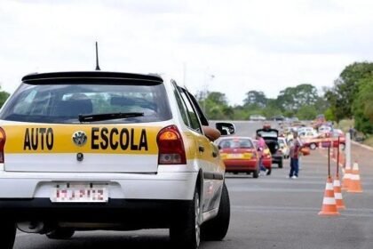 Carro de autoescola em pista de testes do Detran, com cones e outros veículos ao fundo durante exame prático de direção.