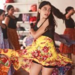 Menina gira com saia colorida durante oficina de dança na Vila das Artes, enquanto outras crianças acompanham a atividade em sala de aula.