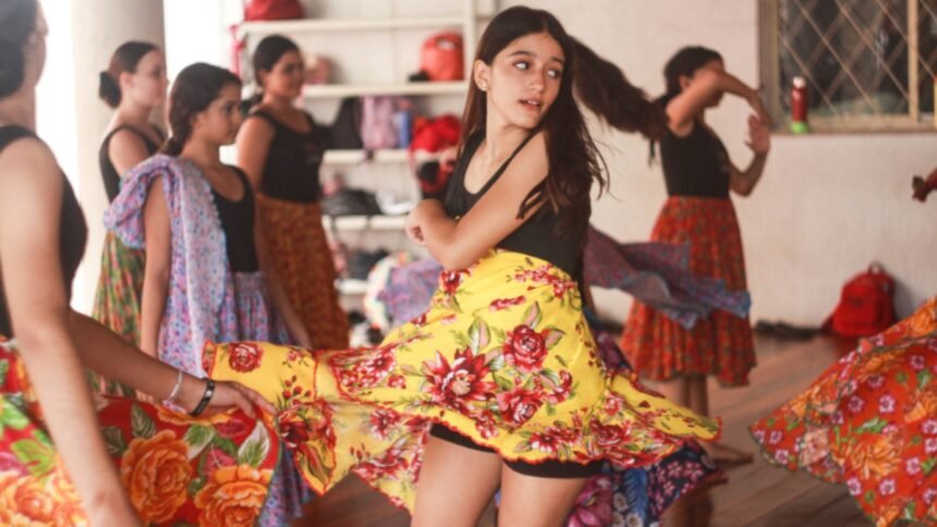 Menina gira com saia colorida durante oficina de dança na Vila das Artes, enquanto outras crianças acompanham a atividade em sala de aula.