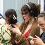 Jovens em frente a um prédio, usando celulares para consultar informações; alguns usam fones de ouvido enquanto observam atentamente a tela.