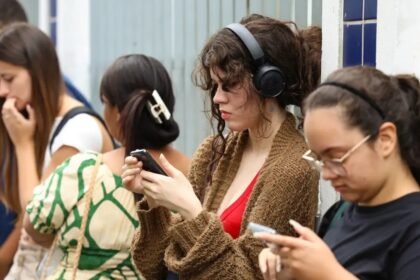 Jovens em frente a um prédio, usando celulares para consultar informações; alguns usam fones de ouvido enquanto observam atentamente a tela.