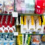 Prateleira de loja com frascos de cola, estojos e diversos materiais escolares organizados em caixas e recipientes transparentes.