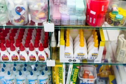 Prateleira de loja com frascos de cola, estojos e diversos materiais escolares organizados em caixas e recipientes transparentes.