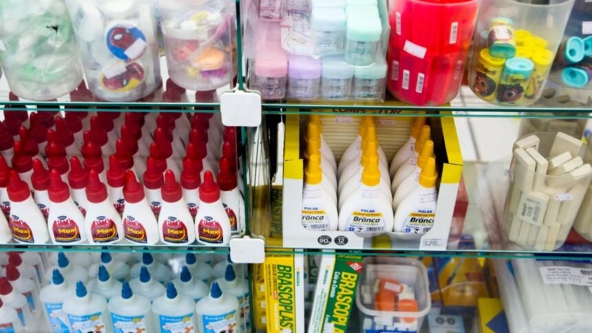 Prateleira de loja com frascos de cola, estojos e diversos materiais escolares organizados em caixas e recipientes transparentes.