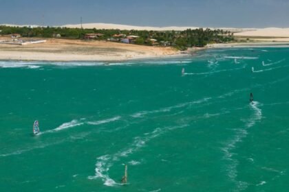 Imagem aérea de uma praia no Ceará com mar em tom esverdeado, dunas ao fundo e várias pessoas praticando windsurf e kitesurf ao longo da costa.