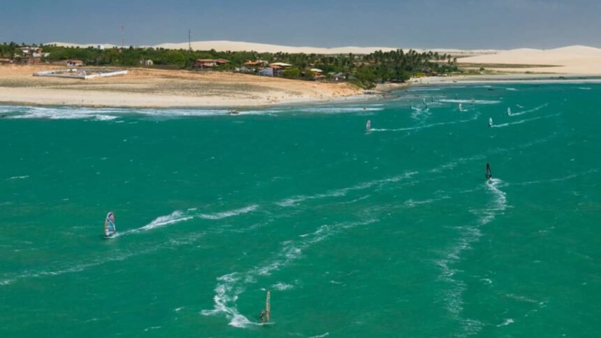 Imagem aérea de uma praia no Ceará com mar em tom esverdeado, dunas ao fundo e várias pessoas praticando windsurf e kitesurf ao longo da costa.