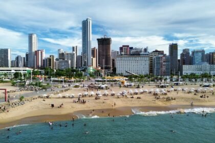 Vista aérea da orla de Fortaleza, com banhistas na praia, guarda-sóis na areia e prédios altos ao fundo, retratando a cidade como destino turístico que movimenta diversos setores da economia.