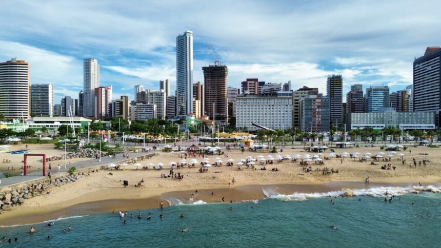 Vista aérea da orla de Fortaleza, com banhistas na praia, guarda-sóis na areia e prédios altos ao fundo, retratando a cidade como destino turístico que movimenta diversos setores da economia.