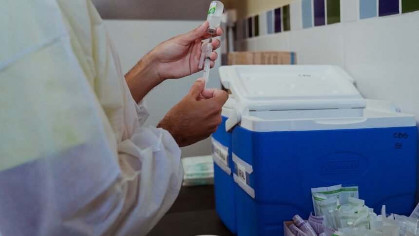 Mãos de profissional de saúde manuseiam seringa e frasco de vacina ao lado de caixa térmica de conservação, em ambiente de posto de vacinação.