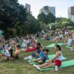 Crianças, jovens e adultos sentados em colchonetes sobre a grama participam de atividades coletivas em área aberta do Parque do Cocó, com prédios ao fundo e clima de convivência familiar.