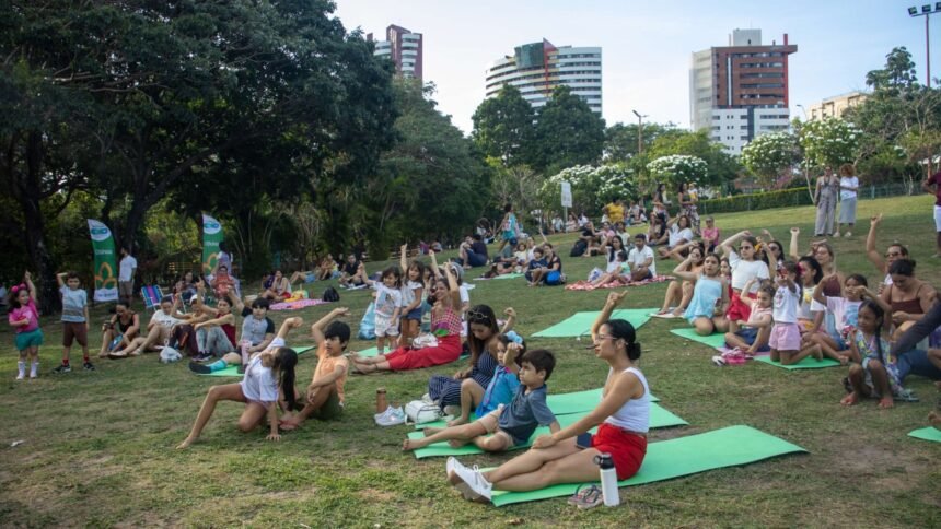 Crianças, jovens e adultos sentados em colchonetes sobre a grama participam de atividades coletivas em área aberta do Parque do Cocó, com prédios ao fundo e clima de convivência familiar.